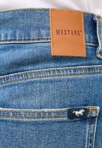 Mėlyni džinsai su rusvu odiniu etiketu, ant kurio užrašyta "MUSTANG", ir maža balta arkliuko emblema ant nugarinės kišenės, puošti geltonomis siūlėmis.