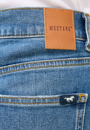 Modré džínsy z denimu s hnedou koženou nášivkou s nápisom "MUSTANG" a malým bielym logom koňa na zadnej vrecku, s žltým prešívaním.
