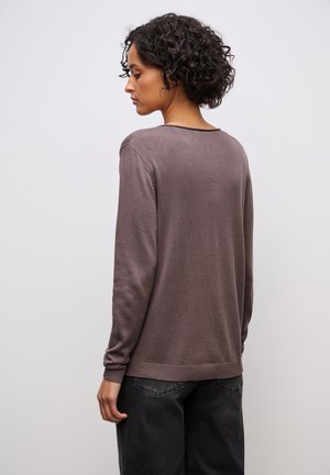 Donna con i capelli corti e ricci, indossa un maglione a maniche lunghe viola e jeans neri, rivolta a sinistra su uno sfondo chiaro e semplice.