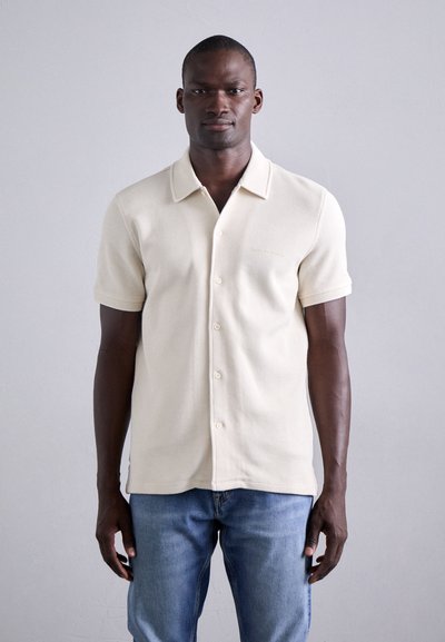 Polo beige à manches courtes avec un motif texturé, col ouvert, et cinq boutons sur le devant, associé à un jean bleu clair.