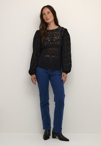 Sort blonde langærmet top med intrikate mønstre og rund hals, parret med blå denimjeans og sorte hælede ankelstøvler.