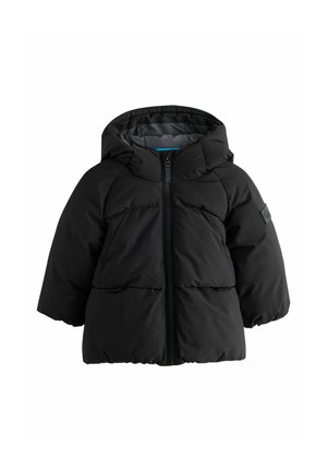 Jong kind draagt een zwarte gewatteerde jas met capuchon en een voorrits en lichtgekleurde broek, staand tegen een effen achtergrond.