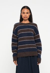 Hosszú, göndör hajú nő barna és sötétkék csíkos bolyhos pulóvert és sötét szoknyát visel, egy sima fehér háttér előtt áll.