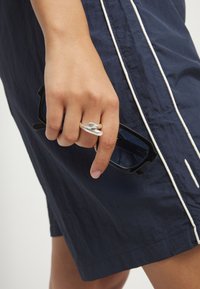 Mano sosteniendo gafas de sol rectangulares negras, con un anillo plateado escultórico, fondo de prenda azul marino con detalle de ribete blanco.