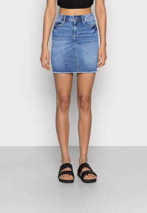 Vrouw die een blauwe denim minirok met een rafelige zoom draagt, een zwart cropped topje en zwarte sandalen met doorgestikte bandjes.