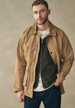 Beige Canvas-Jacke mit einem Cordkragen, ausgestattet mit Druckknöpfen und Seitentaschen, über einem dunkelgrünen Reißverschluss-Pullover und Jeans getragen.