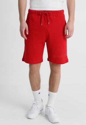 Homme portant un short rouge vif à cordon de serrage, des chaussettes blanches montantes avec un logo bleu, et des baskets blanches de sport, debout devant un fond uni.