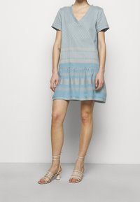 Svetle modré šaty s krátkým rukávem a výstřihem do V, s texturovanými vzory a volánkovým lemem. Nosené s béžovými sandály na platformě, které mají zaoblený podpatek.