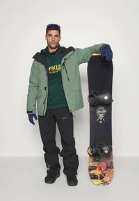 Snowboarder u zelenoj jakni, crnim hlačama i tamnim rukavicama, oslanja se na snowboard s grafičkim dizajnom i crnim vezovima.