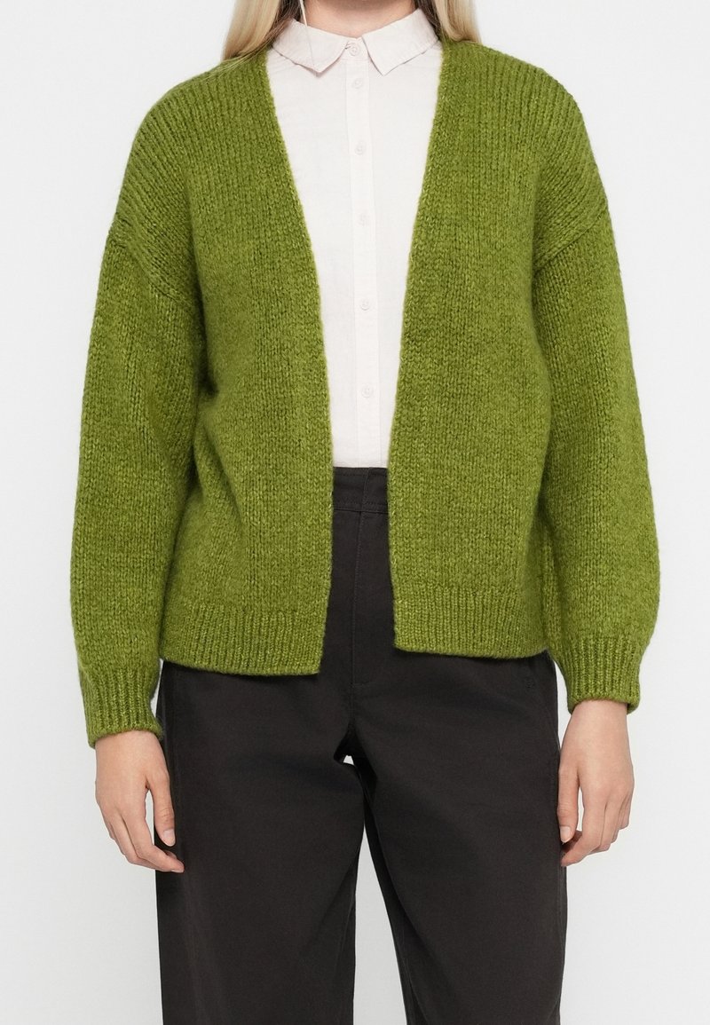 Personne portant un cardigan tricoté vert clair ouvert sur le devant, une chemise blanche boutonnée et un pantalon noir, debout devant un fond uni.