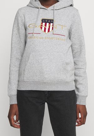 Personne portant un sweat à capuche gris avec une poche avant et le logo "GANT 1949 American Sportswear" en rouge, or et marine sur la poitrine.