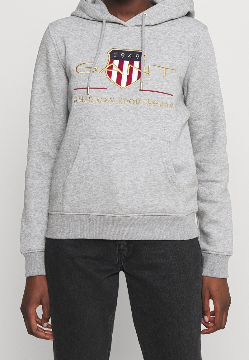 Personne portant un sweat à capuche gris avec une poche avant et le logo "GANT 1949 American Sportswear" en rouge, or et marine sur la poitrine.