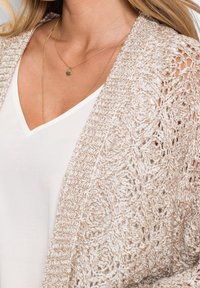 Cardigan beige lavorato a maglia con motivo a pizzo aperto e bordi a coste, indossato su una maglietta bianca con scollo a V. Presenta una delicata collana con pendente dorato.