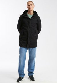 Zwarte lange jas met een capuchon, voorzien van knopen aan de voorkant en twee zijzakken. Gedragen over een lichtblauwe shirt en gecombineerd met een relaxed fit jeans en sneakers.