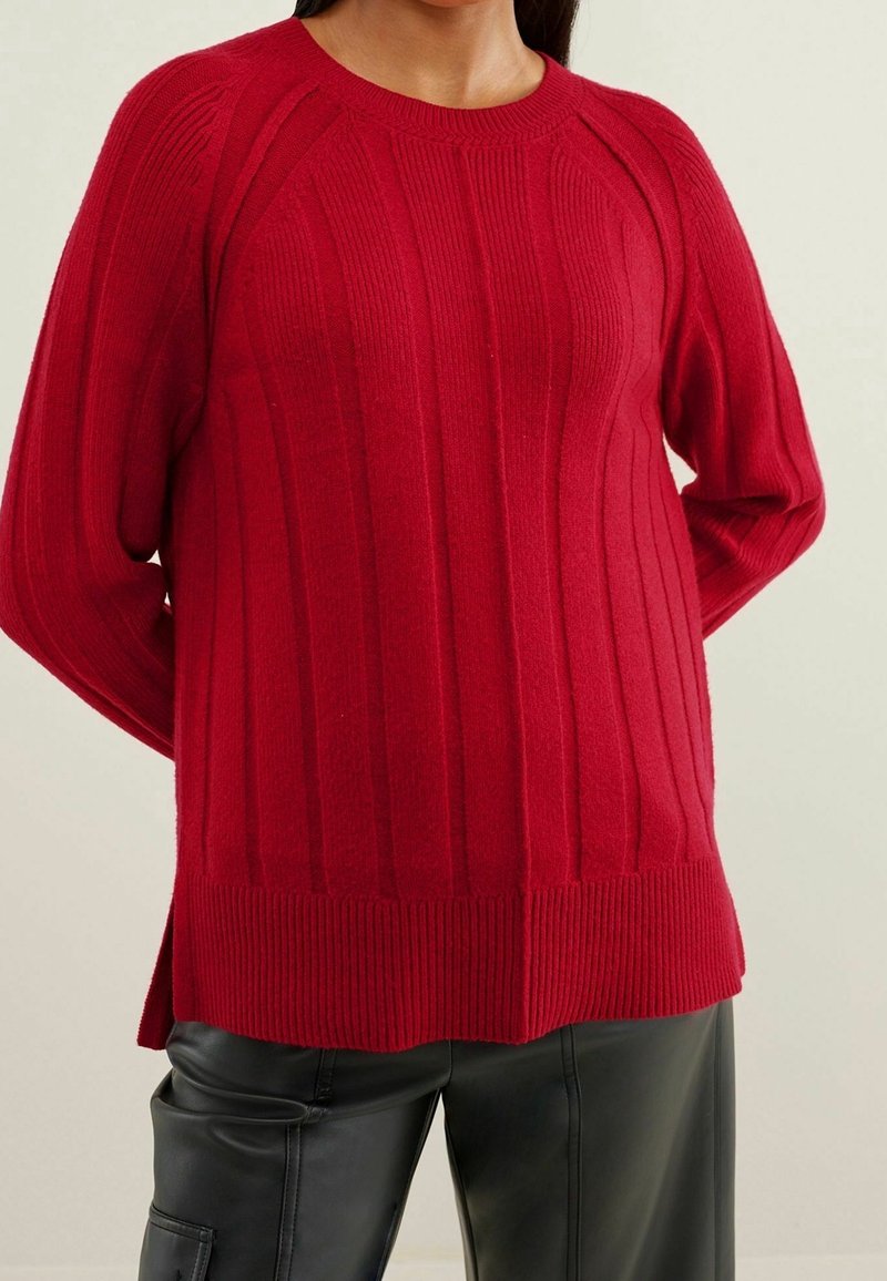 Femme portant un pull en maille rouge à manches longues et un pantalon en cuir noir, debout les mains derrière le dos devant un fond clair uni.