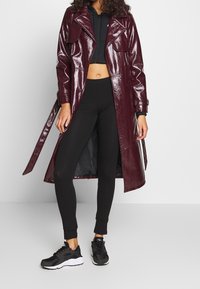 Bordeaux glanzende trenchcoat met twee voorzakken, gecombineerd met een zwart crop top, zwarte leggings en zwarte sneakers.