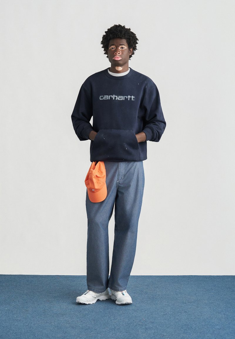 Marineblauer Sweatshirt mit "Carhartt"-Logo, Jeanshose, orangefarbene Kappe an der Taille befestigt, weiße Turnschuhe. Einfaches Design und lässige Passform.