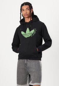 Svart hoodie med en pixelerad grön Adidas-logga, dragsko för huvan och framficka. Bärs med grå denimshorts.