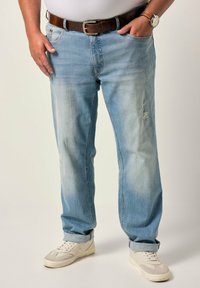 Jeans de denim de color azul claro con un corte relajado, que presentan áreas descoloridas y detalles deshilachados, combinados con zapatillas blancas. Un cinturón de cuero marrón acentúa el conjunto.