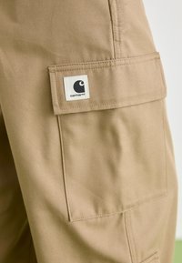 Tan katoenen cargobroek met een grote zijzak en een vierkant label met een zwart logo op de linkerdij. Duurzaam materiaal met een gladde textuur.