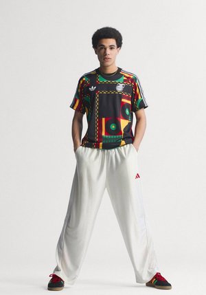 Jeune homme debout les mains dans les poches de son pantalon blanc, portant un t-shirt Adidas à motifs noirs, rouges, jaunes et verts, ainsi que des baskets noires avec des lacets rouges.