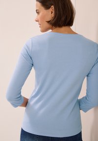 Haut en coton bleu clair, à manches longues, avec un col rond, une silhouette ajustée et une texture lisse, associé à un jean bleu.