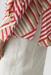 Person trägt ein langärmliges, rot-beige-weiß gestreiftes Hemd mit weißen Knöpfen und eine cremefarbene, strukturierte Hose, Hand an der Seite.