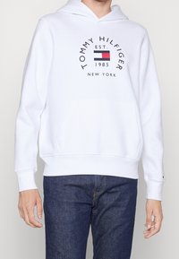 Sudadera blanca con capucha que presenta un bolsillo frontal, puños y dobladillo acanalados, y un logo central con texto y un diseño de bandera de colores.
