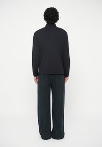 Pessoa com cabelo encaracolado de costas, vestindo uma blusa preta de mangas compridas e calças de ganga escuras de perna larga, contra um fundo simples.