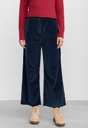 Person iført marineblå vidde fløjlsbukser, rød langærmet top og beige snørestøvler ved anklen, stående på gråt gulv.