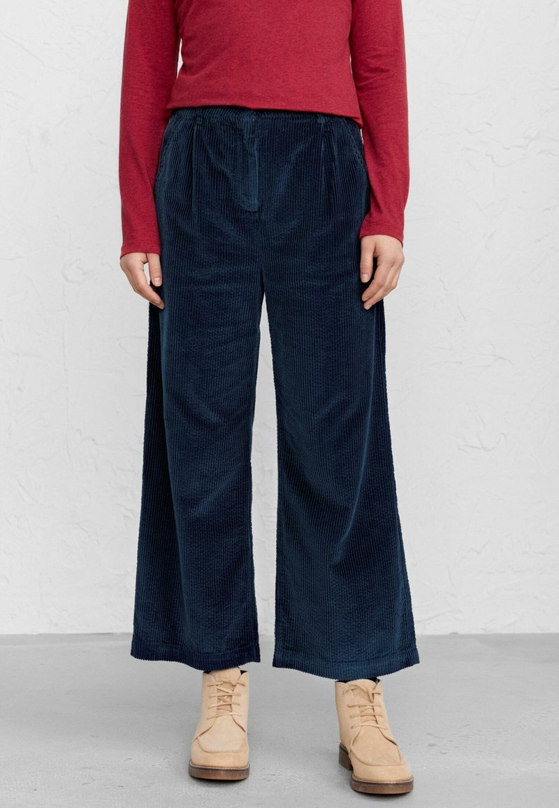 Personne portant un pantalon large en velours côtelé bleu marine, un haut à manches longues rouge, et des bottines beige à lacets, debout sur un sol gris.