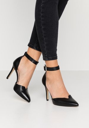 Personne portant des escarpins noirs à bout pointu avec des brides de cheville et un jean skinny foncé, se tenant sur un sol blanc.