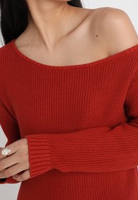 Roter, gestrickter Pullover mit One-Shoulder-Design, gerippten Bündchen und strukturiertem Stoff. Keine sichtbaren Muster oder Akzente.