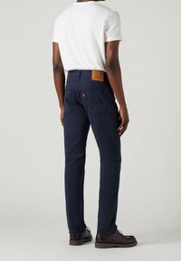 Pantaloni dritti blu navy con una pettorina in pelle marrone testurizzata sul retro della vita e due tasche posteriori. Abbinati a una maglietta bianca.