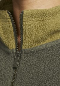 Fleecejacka i olivgrön och grå, med hög krage, hel dragkedja, mjuk textur och framträdande sömdetalj.