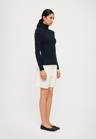 Jūrinės mėlynos ilgomis rankovėmis turtlenecko viršus, derinamas su kreminės spalvos šortais ir juodais butais. Lygus audinys, prigludęs dizainas ir minimalistinė stilius.