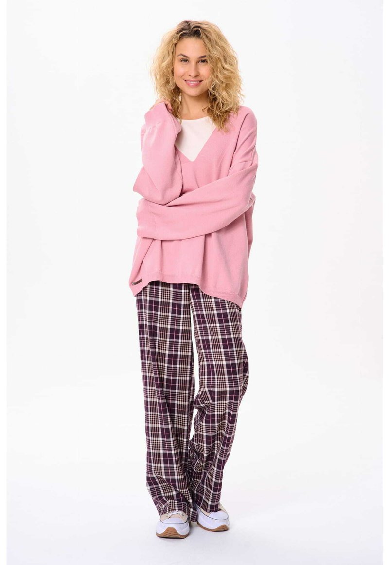 Helle rosa, übergroße Sweater mit V-Ausschnitt, kombiniert mit lässigen, karierten Schlafanzughosen in dunklem Purpur, Weiß und grünen Farbtönen. Weiße Sneakers runden den Look ab.