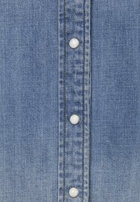 Tissu en denim bleu clair présentant une rangée verticale de cinq boutons-pression argentés avec des accents blancs, une texture lisse et des détails de couture subtils.