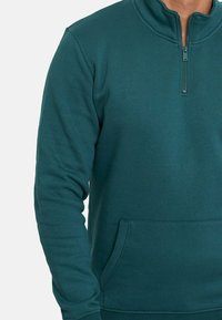 Pólder de forro polar verde con cierre de cremallera de un cuarto, puños acanalados y un bolsillo tipo canguro en la parte frontal. Textura suave, diseño casual y ligero.