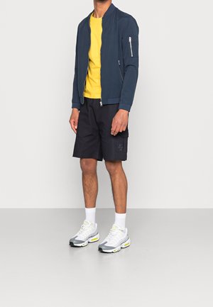 Marine bomberjakke, gul t-shirt, sorte cargoshorts og grå sneakers med gule detaljer. Jakken har en lynlåslomme på armen.