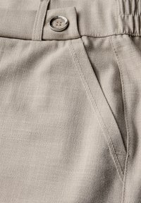 Pantalons beiges en tissu texturé, avec une poche avant et un bouton en argent brillant à surface lisse.