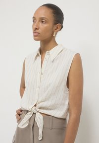 Camicia bianca senza maniche, con colletto e righe blu, annodata in vita. Realizzata in tessuto leggero, presenta chiusura con bottoni e una vestibilità rilassata.
