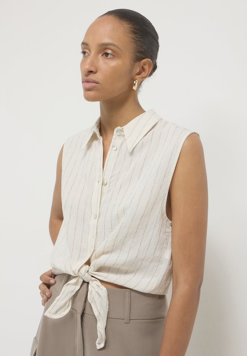 Camicia bianca senza maniche, con colletto e righe blu, annodata in vita. Realizzata in tessuto leggero, presenta chiusura con bottoni e una vestibilità rilassata.