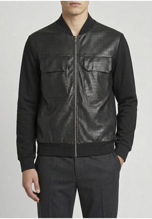 Hombre con chaqueta negra con cremallera, con delante de piel sintética texturizada y bolsillos con solapa, combinada con pantalones gris oscuro.