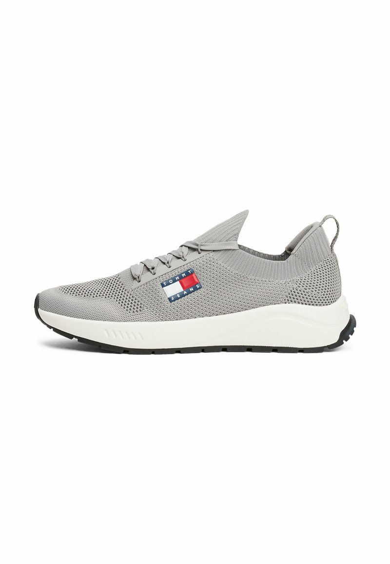 Scarpa sportiva grigia con tomaia in rete, intersuola bianca e suola nera. Presenta un'etichetta con logo con elementi rossi, bianchi e blu.