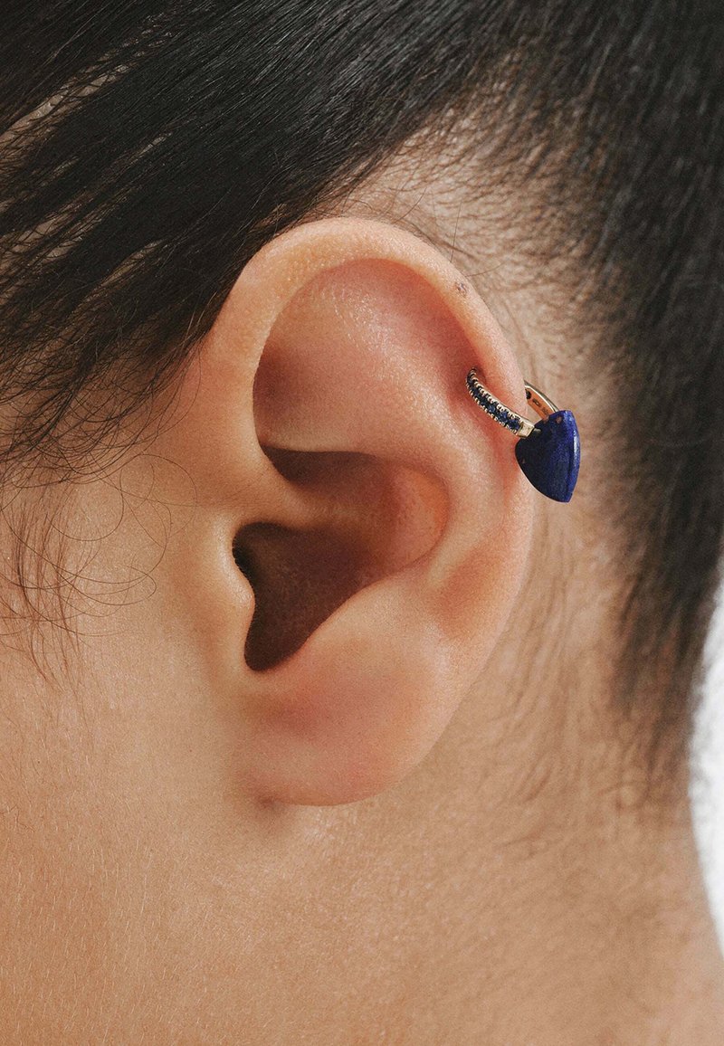Pendiente de aro dorado sujeto a la parte superior de la oreja, que presenta una piedra en forma de corazón de color azul profundo y pequeñas gemas a lo largo del borde del aro.
