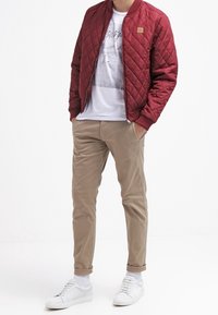 Vinröd quiltad bomberjacka, vit t-shirt med tryck, beige slim fit-byxor och vita sneakers. Enkel design med ribbstickade muddar och fåll.