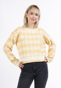 Sárga és fehér mintás, rövidített pulóver kerek nyakkivágással és bordás mandzsettával, fekete farmernadrággal viselve. Az anyag lágy és textúrált.