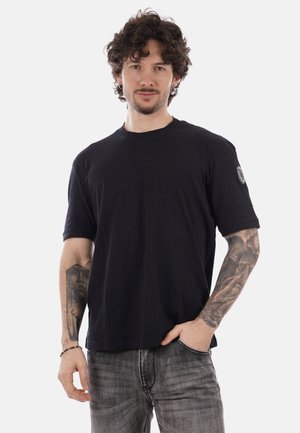 Uomo con capelli ricci che indossa una maglietta nera e jeans grigi, con tatuaggi dettagliati su entrambi gli avambracci, in piedi contro uno sfondo semplice.