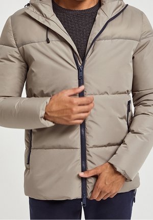 Mann trägt beige gesteppte Jacke mit schwarzem Reißverschluss, die er über einem dunklen Strickpullover und dunklen Hosen verschließt.